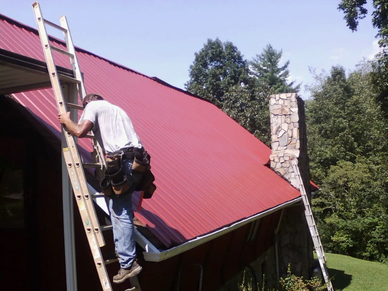 Roofer on a ladder beside a red metal roof and stone chimney — Soffit & Fascia Repair by Roofing Brothers in Norlina